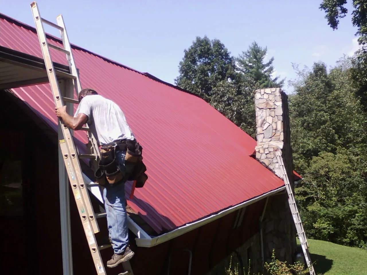 Roofer on a ladder beside a red metal roof and stone chimney — Soffit & Fascia Repair by Roofing Brothers in Paw Paw